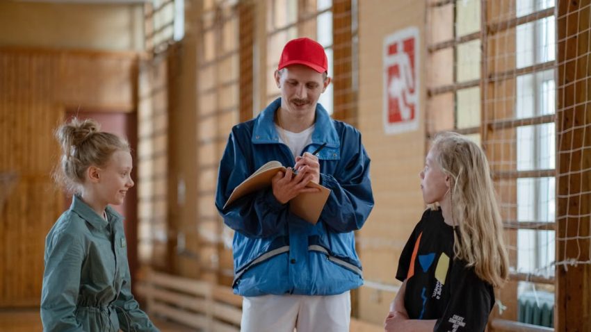 A male coach interacts with two girls during a sports activity in an indoor gym.